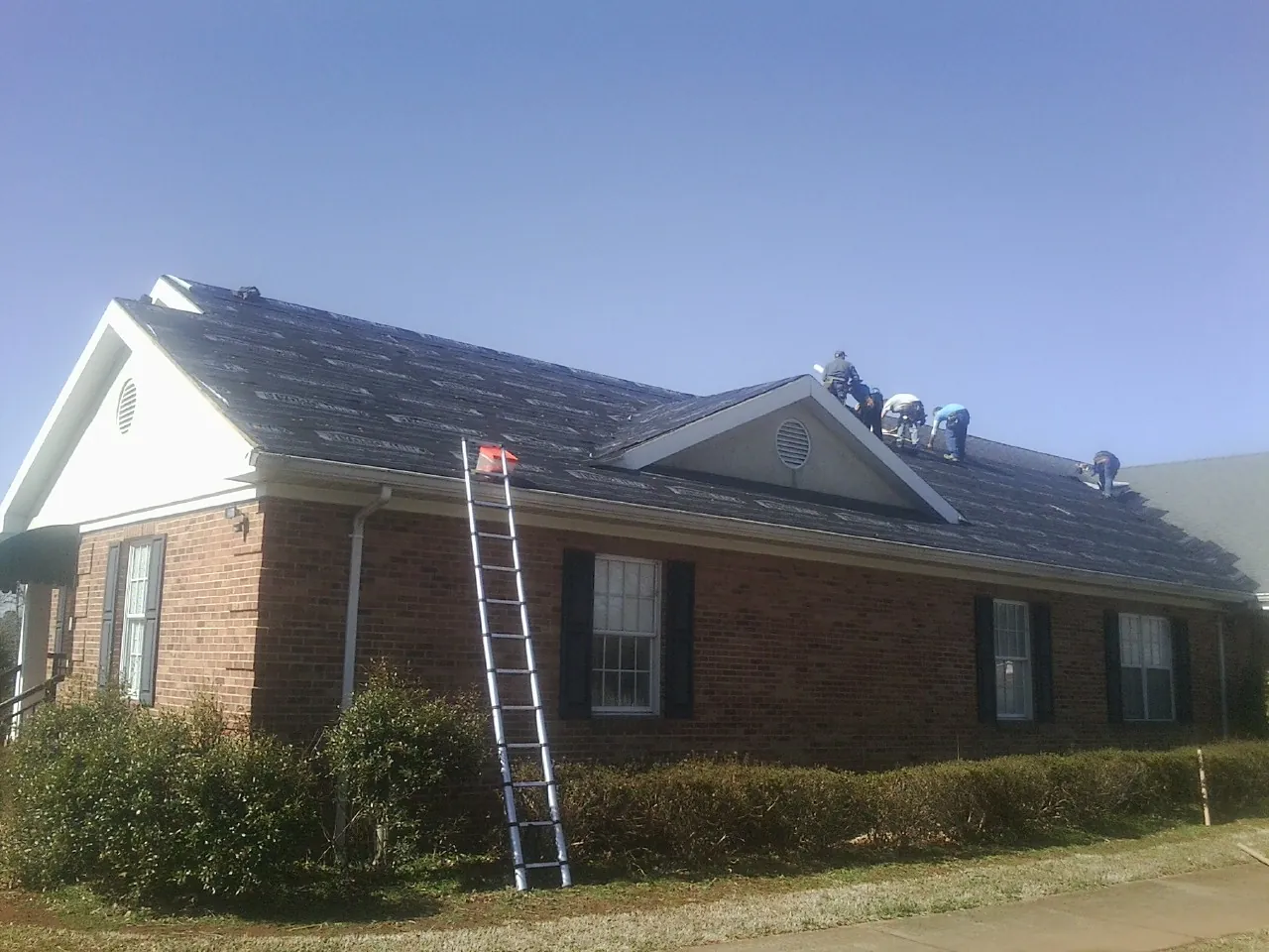 Crew tearing shingles off a brick ranch roof with a ladder set — Emergency Roof Repair work by our crew in Bushwood