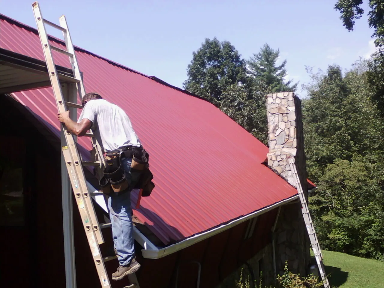 Roofer on a ladder beside a red metal roof and stone chimney — Soffit & Fascia Repair by Roofing Brothers in Bushwood